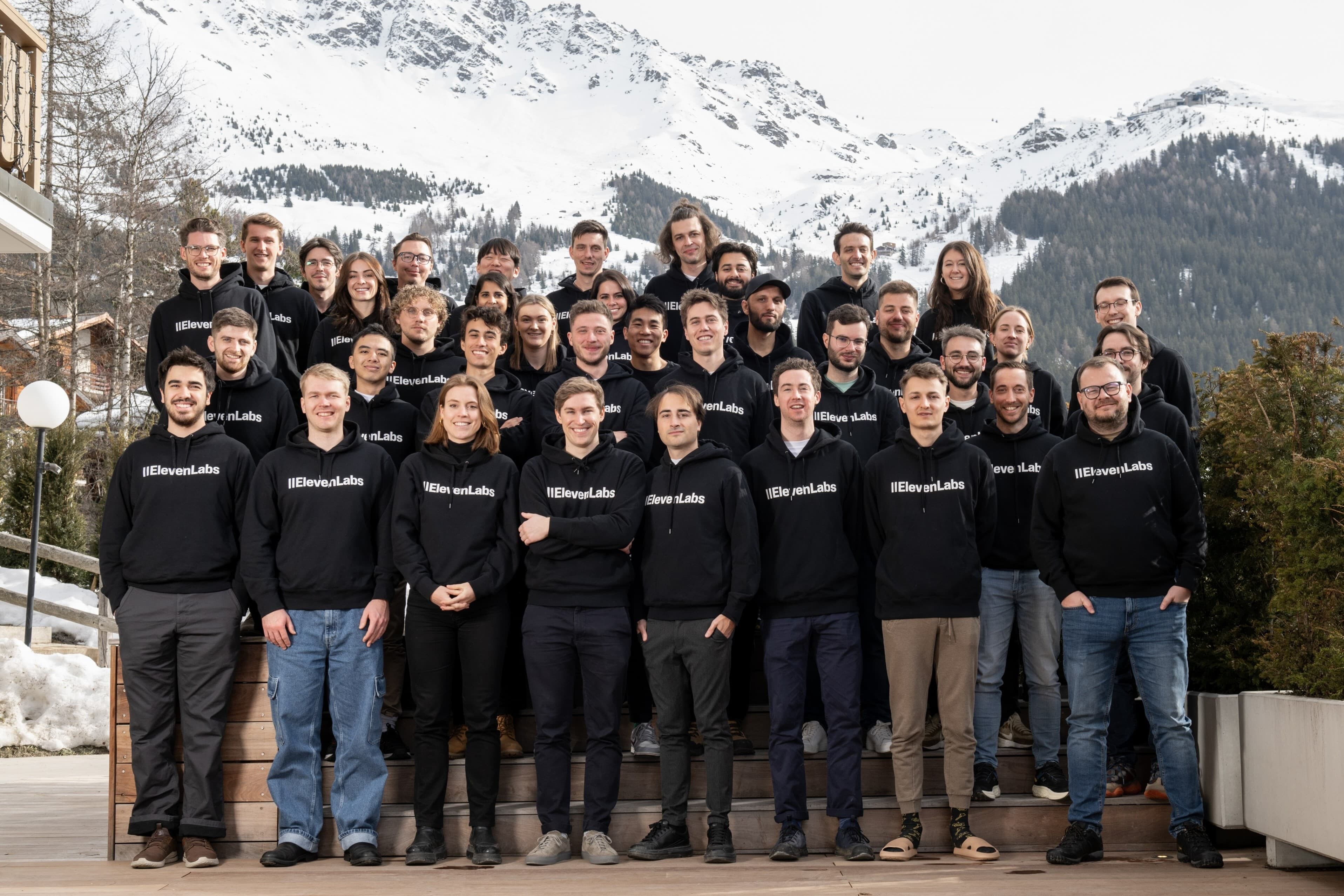 Group photo of people wearing black hoodies with "IIEevenLabs" printed on them, outdoors with snow-covered mountains in the background.